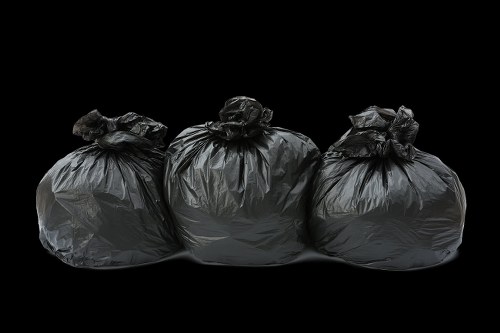 The image displays three black plastic rubbish bags filled with waste, placed in a line on a dark surface against a black background. The bags appear to be made of thick, slightly glossy material, showing some creases and folds on their surfaces. They are gathered at the top and tied off, with the knots visible, indicating they are sealed for disposal. The bags vary slightly in shape and size, with some bulging more prominently, suggesting different contents. The environment appears to be a controlled, indoors setting or a shaded outdoor area with minimal ambient light focused on the bags, emphasizing their texture and form. This visual could relate to rubbish collection or disposal services offered by offices or commercial premises, such as those provided by Office Clearance Whitechapel, highlighting the common appearance of waste packaging ready for removal or recycling in a typical urban setting near Whitechapel or nearby zones in East London. The professional presentation underscores the importance of proper waste management and clearance services tailored to local needs.