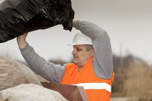 A professional waste removal worker, wearing a white safety helmet and an orange high-visibility vest over a grey long-sleeve shirt, is lifting a large black plastic rubbish bag. The worker is positioned outdoors on a construction or clearance site, with a blurred background that suggests an overcast sky and open land. The individual is focused on lifting the bag, which appears full and heavy, with its glossy surface reflecting some light. Nearby, there are other materials possibly related to rubbish or debris, including a large beige sack and a piece of reddish-brown material, indicating ongoing clearance activity. This scene aligns with rubbish removal services offered by companies like Office Clearance Whitechapel, based near the Tower Hamlets area, serving Whitechapel and surrounding districts, with a focus on professional waste collection and disposal in urban or semi-urban environments.