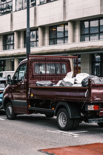 A small, dark red flatbed pickup truck parked on a city street adjacent to a modern, multi-story office building with large tinted windows and concrete exterior walls. The truck's cargo area is loaded