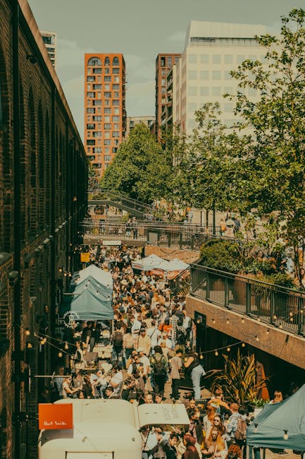 The image shows a busy outdoor market stall located under a yellow canopy, with people shopping and browsing. In the foreground, there are various fresh vegetables and produce, including green broccol
