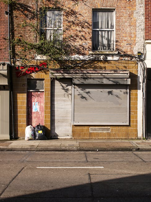 A narrow urban alleyway with uneven, muddy ground and patches of dampness. On the right side, there are various discarded items including a large white washing machine, a wooden crate or box, and a pa