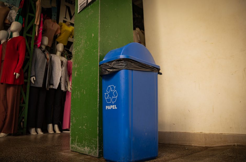 A black wheelie bin with white lettering labeled 'COMMERCIAL WASTE ONLY' is positioned in front of a storefront with large glass windows and wood-paneled exterior painted in a deep red color. The bin